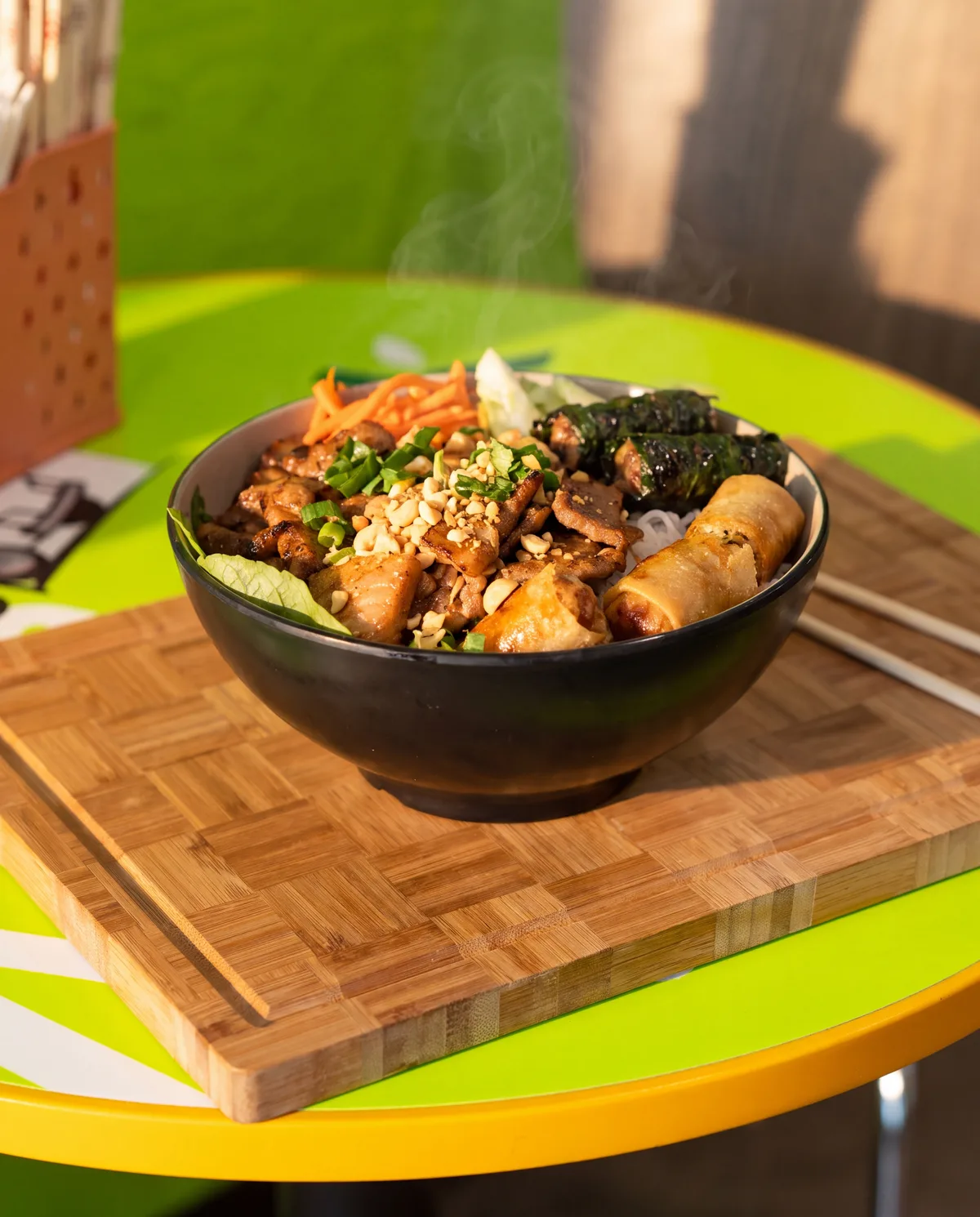 Our Bún Đặc Biệt on a bamboo board on the green table: grilled pork, betel leaf beef, spring roll, herbs, pickles and peanuts, fish sauce on the side
